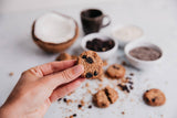 Hand holding a cookie with ingredients like coconut, chocolate, and spices on a light surface.