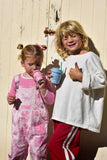Two young girls standing outdoors in front of a weathered cream-colored wooden door. The girl on the left wears pink overalls over a pink shirt and holds a pink cup.