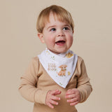 A smiling baby wearing a beige ribbed long-sleeve top and a white triangular bib printed with a cute brown lamb, small flowers, and a picket fence, photographed against a solid beige background.