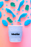 Blue cup with 'bbeeino' branding on a pink background with blue and green abstract shapes.