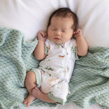 A sleeping newborn baby wearing a white one-piece bodysuit with a woodland animal print and sage green trim, lying on a textured mint green blanket on a white bed.