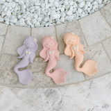 Three pastel-colored mermaid toys on a stone surface with pebbles in the background.