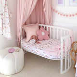 A white wooden baby crib with a pink canopy, decorated with a pink floral mattress, a pink flower-shaped pillow, and a doll, set in a soft pink nursery.