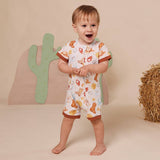 A toddler stands barefoot in a studio setting wearing a short-sleeve one-piece romper with a western food print. The romper has a white base with orange, yellow, and brown illustrations of tacos, burritos, and hot dogs, plus brown trim on the sleeves and legs.