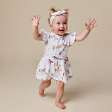 A baby girl wearing a white short-sleeve dress with a desert animal print and a matching bow headband, captured mid-step against a solid beige studio background.