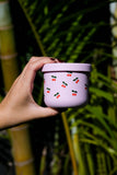 Hand holding a pink container with cherry pattern against a green leafy background