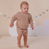 Baby in a beige knit outfit standing against a plain background
