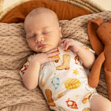 A sleeping newborn baby wearing a white short-sleeve bodysuit with a colorful cowboy boot and western-themed print, lying on a textured beige blanket next to a brown corduroy bunny toy.