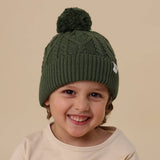 Child wearing a green knit beanie with a pom-pom on a beige background
