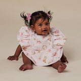 A baby wearing a white ruffled bib with a pink and gold western-themed print, sitting on a beige studio background.
