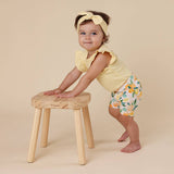 Baby in yellow top and floral shorts standing on a wooden stool against a beige background