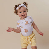 A toddler girl wearing a white short-sleeve henley shirt with a cute woodland animal print, yellow shorts, and a matching white bow headband, posing against a solid beige background.