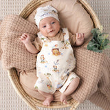 A baby lying on their back in a woven basket, wearing a cream-colored romper and matching hat with a whimsical pirate-themed print. The baby is surrounded by cozy beige blankets.