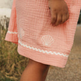 A close-up of a child wearing a peach pink gauze cotton dress or romper with white scallop trim and embroidered white seashells.