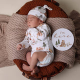 Newborn baby in a basket wearing a white outfit with bear patterns, surrounded by brown and beige textures.
