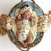 Newborn baby in a floral outfit and headband, wrapped in a yellow blanket, lying on a woven basket with a white wooden background.