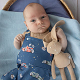 Baby in a blue outfit with space-themed design, holding a beige plush toy, sitting in a wicker chair with a blue cushion.