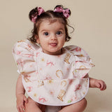 A baby wearing a light pink bib with a western-themed print of boots, horseshoes, bows, and flowers, styled with pink hair bows against a solid beige background.