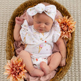 A sleeping newborn baby wearing a white short-sleeve bodysuit with a cute ice cream cone print and a matching bow headband, posed inside a woven basket with pink fabric lining and decorative flowers.