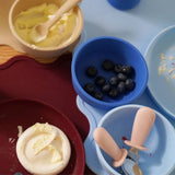 Assorted baby feeding items including bowls with food and utensils on a blue background