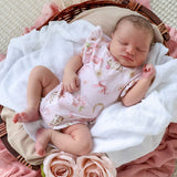 A sleeping newborn baby wearing a light pink short-sleeve romper with a floral and horseshoe pattern, lying in a wicker basket lined with white and pink blankets.