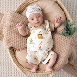 A baby lying on their back inside a woven rattan bassinet, wearing a cream-colored pirate-themed romper and matching hat. The romper features colorful illustrations of a treasure chest, a pirate, and a ship's wheel, and is paired with a matching beige t-shirt.