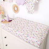 A floral baby changing pad on a white changing table in a nursery setting, with baby clothes hanging on the wall and small decorative items nearby.