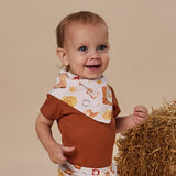 A smiling baby wearing a rust brown short-sleeve romper and a white bib with cowboy boots, stars, and a guitar print, standing next to a bale of hay against a beige studio background.