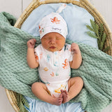 A sleeping newborn baby is lying in a woven bassinet, wrapped in a mint green textured blanket and light blue fabric. The baby is wearing a white short-sleeve bodysuit and matching beanie hat with a colorful octopus and sea creature print.