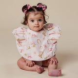 A baby girl wearing a ruffled bib with a western boot and horseshoe pattern, sitting on a beige studio background.