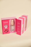 A baby bottle with a pink cap presented in a pink gift box, with a second matching box standing beside it on a warm beige studio background.