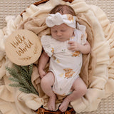 A sleeping newborn baby wearing a white ruffled bodysuit with a cute animal print and a matching bow headband, lying in a wicker basket lined with cream knit blankets. A round wooden sign reading 'Hello World' is next to the baby.