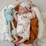 A sleeping newborn baby wearing a cream-colored zip-up romper and matching hat with a vintage car pattern, nestled in a brown blanket next to a mint green plush elephant toy on a textured cream blanket.