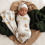 A sleeping newborn baby swaddled in a white wrap blanket with a brown bear and bunny print, wearing a matching beanie hat. The set is styled on a white surface with tan fabric and a dark green chunky knit blanket.