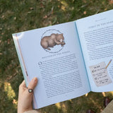 Open book with a bear illustration and text held by a hand outdoors.