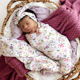 A sleeping newborn baby wrapped in a white floral swaddle blanket and matching floral headband, resting inside a woven basket with purple knit blankets.