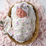 A sleeping newborn baby swaddled in a white muslin blanket with a colorful floral print, wearing a matching bow headband, resting inside a woven wicker basket on a pink blanket.