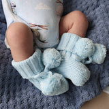 Baby wearing light blue knitted socks with pom-poms on a textured gray blanket.