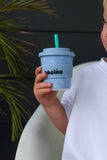 Child holding a blue cup with a straw, against a dark background with greenery.