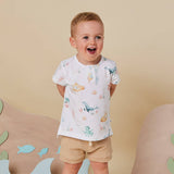 A young toddler boy wearing a white short-sleeve t-shirt with a colorful sea-creature print and beige shorts, standing against a warm beige background with decorative wooden shapes.