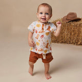 A smiling baby wearing a short-sleeve western-themed t-shirt and brown shorts, standing barefoot in front of a beige studio backdrop with a hay bale and cowboy hat prop.