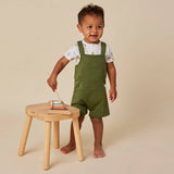 A toddler stands barefoot next to a small wooden stool, wearing green overall shorts over a white patterned t-shirt, against a solid beige background.