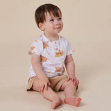 A baby sitting on a beige studio floor wearing a white short-sleeve top with a brown animal print and tan shorts.