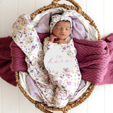 A sleeping newborn baby is swaddled in a floral footed swaddle blanket and wearing a matching floral bonnet, resting in a woven basket with purple knit blankets and a white 'I'm here' sign.