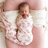 A sleeping newborn baby swaddled in a light pink swaddle blanket with a western print of boots, horseshoes, bows, and stars, resting on a pink pillow with a cream knit blanket.