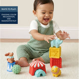 A baby sitting on a light beige carpet plays with colorful circus-themed wooden blocks and animal toys. The baby wears a white t-shirt and a sage green knit overalls.