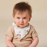 A baby wearing a beige short-sleeve bodysuit and a white printed bandana bib with illustrations of a pirate and palm trees, photographed against a solid beige background.