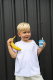 A young blonde boy wearing a white t-shirt and light gray sweatpants stands in front of a dark gray corrugated metal wall, holding a yellow banana in one hand and a blue striped cup with a straw in the other.