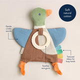 A duck-shaped soft baby teething toy with blue wings, a green head, orange beak and feet, and a brown body. The toy has callout labels pointing to features like gentle crinkle sounds, textured ribbon, and soothing sore gums, plus a dark blue circle that reads 'Soft natural cotton' on a light beige background.