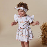 A toddler girl wearing a white ruffle-sleeve dress and matching headband with a desert horse print, standing next to a bale of hay against a beige studio background.